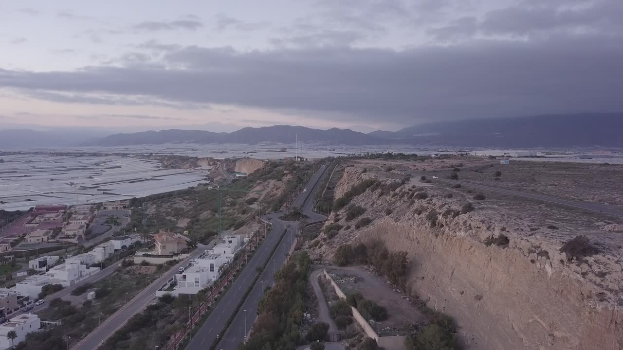 Aerial view of a highway winding through a landscape with mountains, greenhouses, and a residential area