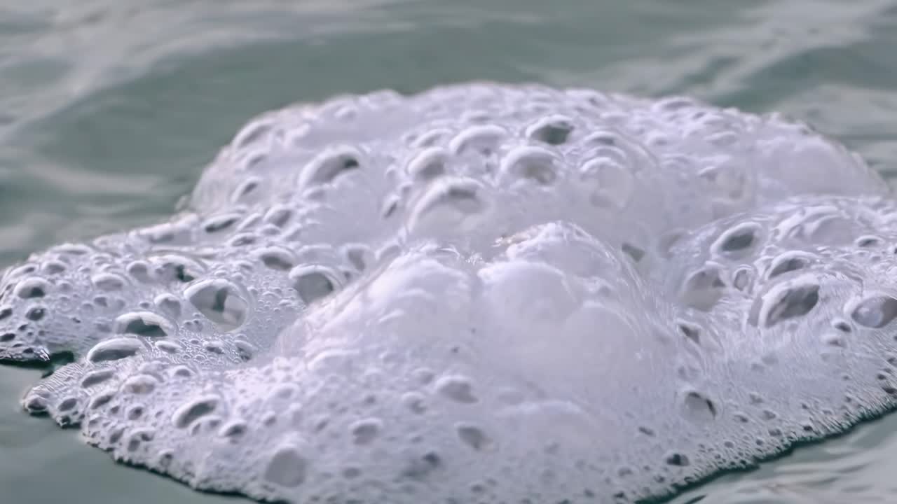 Close-up video of white sea foam bubbles on water surface, captured from a low angle