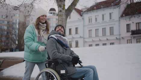 happy caucasian woman taking her disabled friend in wheelchair for a walk in the winter city while talking and laughing together