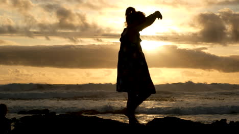 free-spirited girl in dress swirls around, silhouette against gold ocean sunset