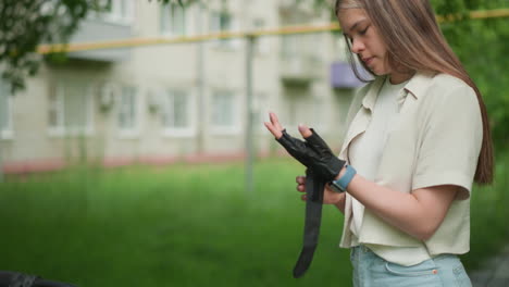 young woman stands outdoors puts on biker gloves, readying for an active day, background includes lush greenery, trees, and a blurred view of residential buildings