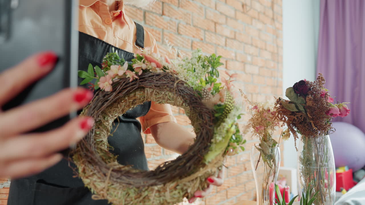 Female florist wearing black apron holding floral wreath decorated with green leaves and pink blossoms, taking selfie with smartphone in rustic workshop against brick wall, handmade decoration moment