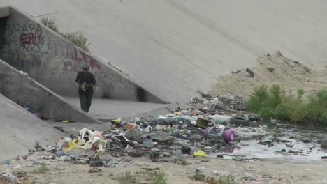 a man walks amongst garbage and litter in a drainage ditch