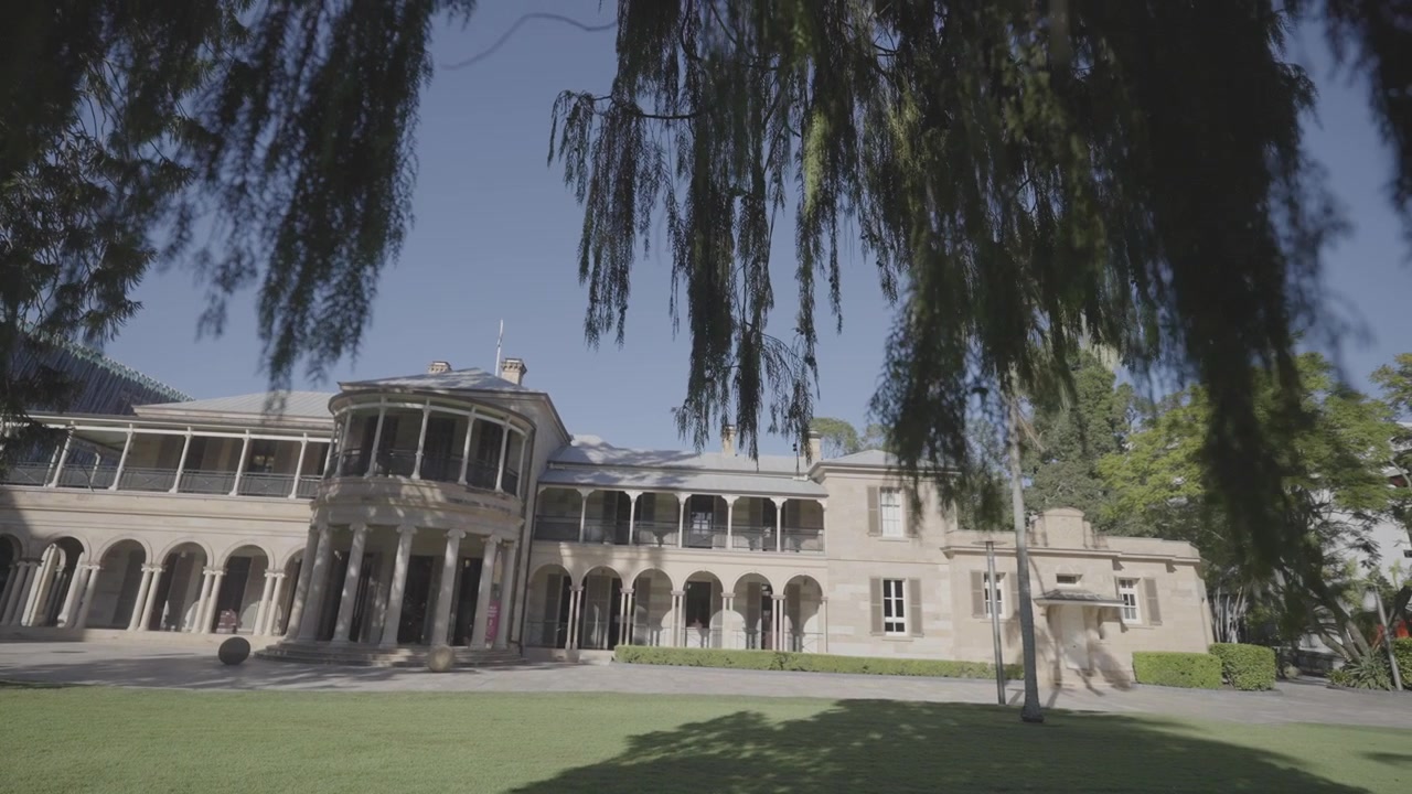 A wide view shows a heritage sandstone building with a colonnaded verandah, city high rise towers rising behind it.