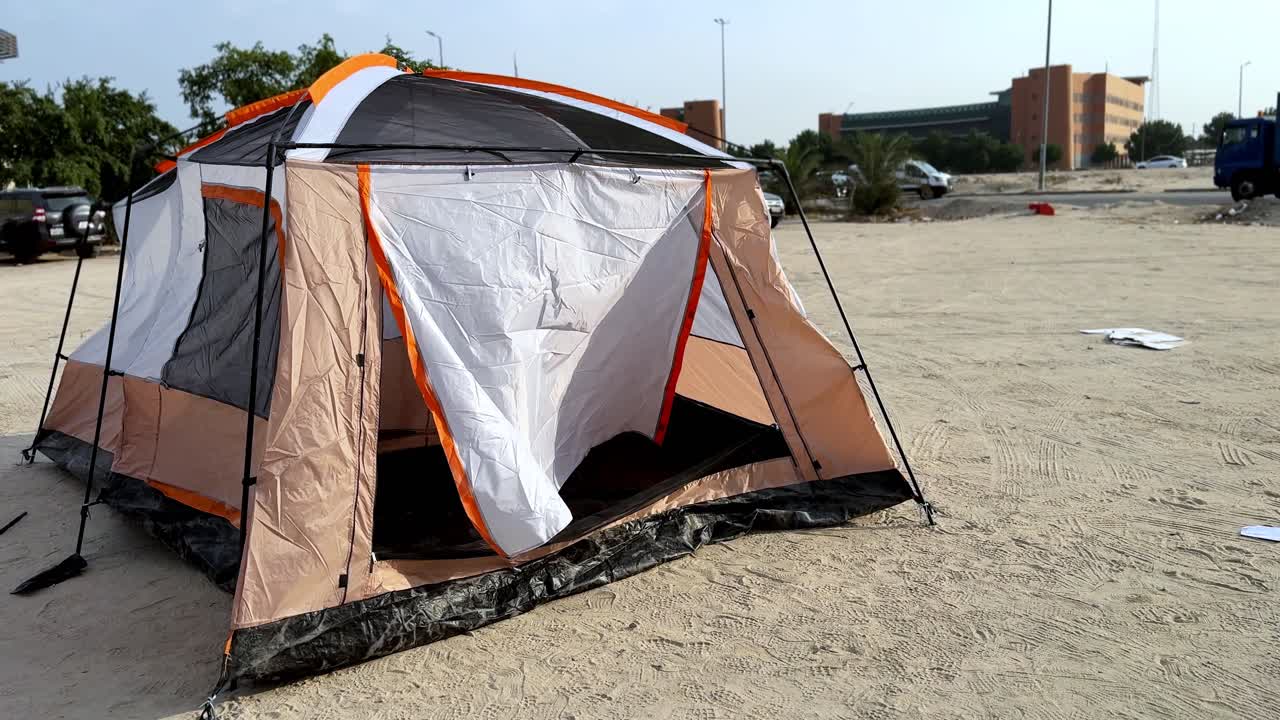 Daytime wide of a pitched camping tent with open door on a sandy lot near a road; cars, trees, and buildings in background, clear sky, no people—great for outdoor gear or urban camping B-roll