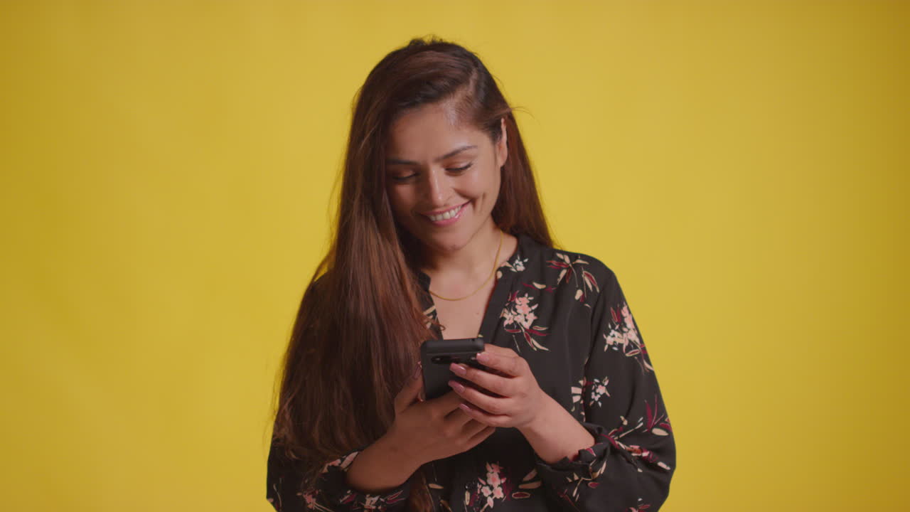 Scatto in studio di donna sorridente con telefono cellulare che riceve buone notizie su sfondo giallo