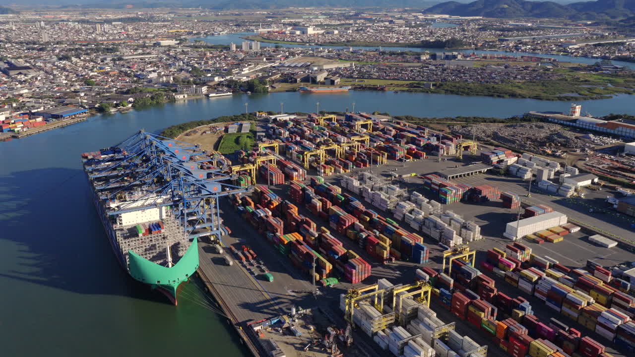 Ascending drone captures container vessel loading at Port of Itajaí with cranes and city during sunny afternoon