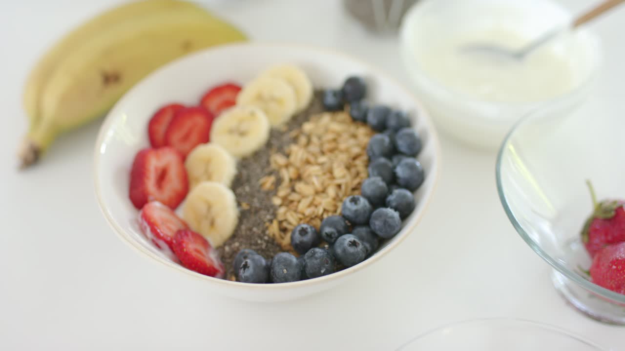 La telecamera si allontana per rivelare una ciotola bianca con frutta, una striscia di chia, yogurt e banane su un piano di lavoro.