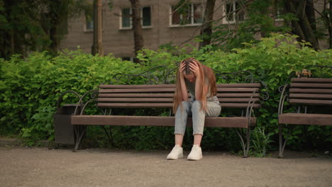 woman sits on wooden bench, in somber posture, her long hair cascades down as she supports her head with her hands, surrounded by lush greenery, trees, and an urban building in background