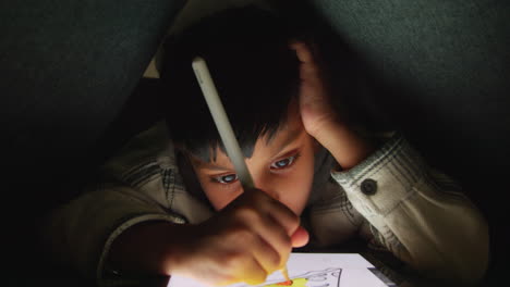 close up of young boy in home made camp made from cushions playing and drawing with digital tablet and stylus at night 2