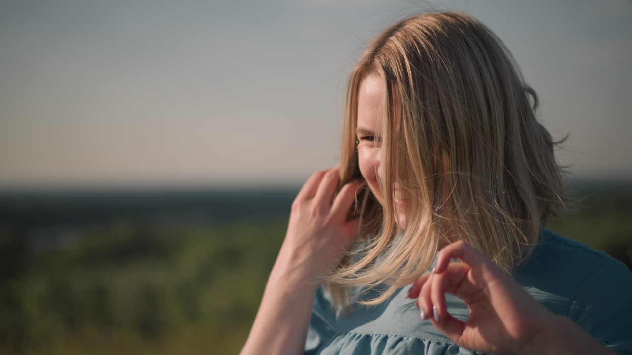 A close-up shot of a woman in a blue shirt as the wind blows her hair. She gently moves her hair away from her face, creating a natural and serene moment with a blurred background