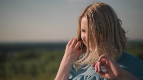 a close-up shot of a woman in a blue shirt as the wind blows her hair. she gently moves her hair away from her face, creating a natural and serene moment with a blurred background