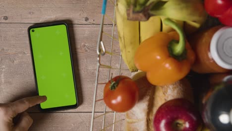 foto de estudio superior de una persona que usa un teléfono móvil de pantalla verde junto a alimentos básicos en la cesta de la compra de alambre del supermercado 2