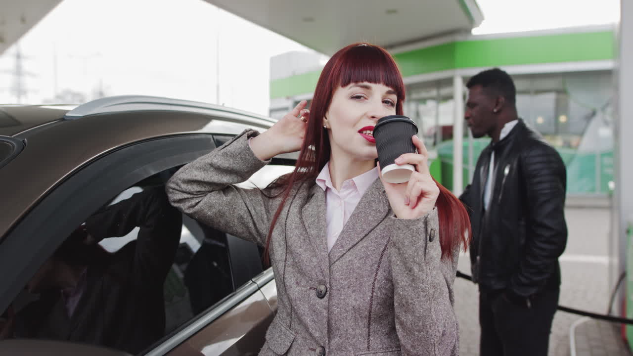 Woman drinking coffee at gas station while man pumps gas
