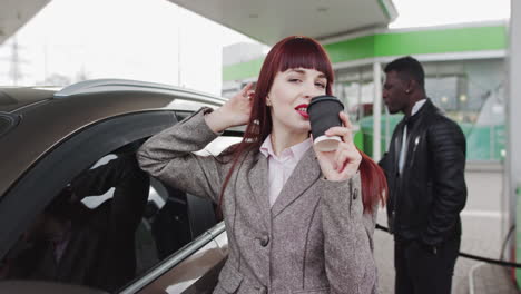 woman drinking coffee at gas station while man pumps gas