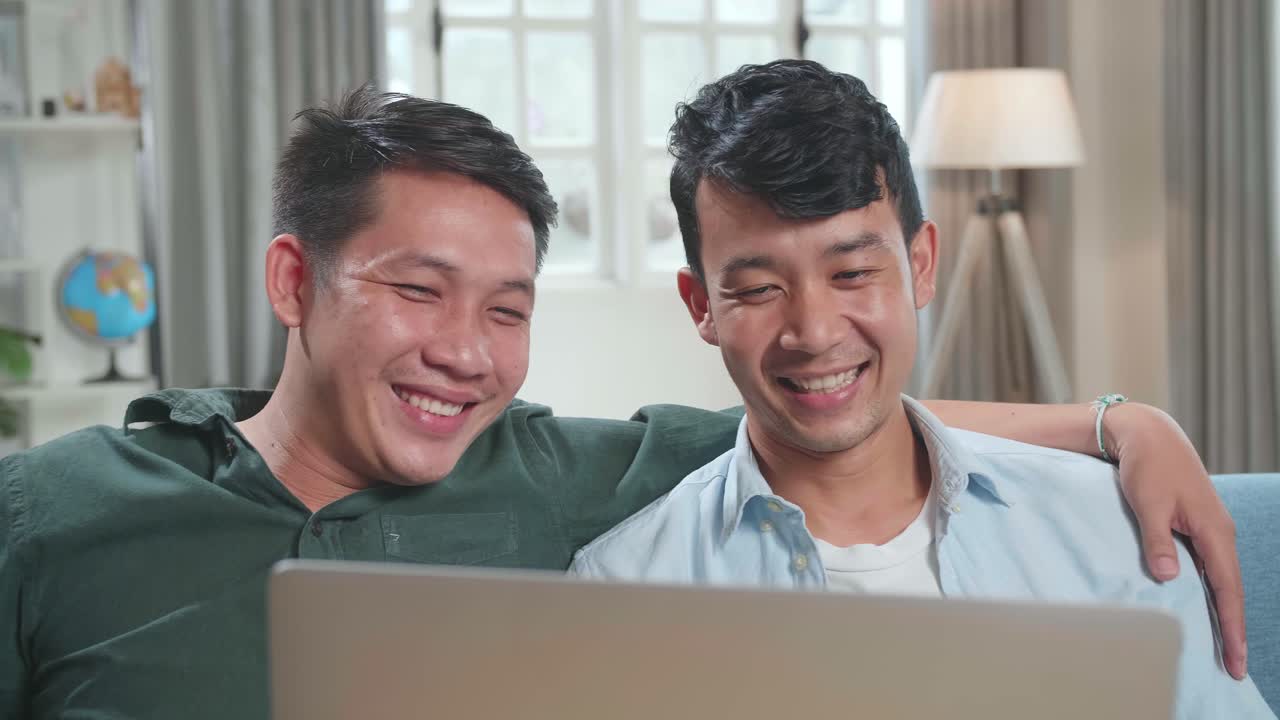 Portrait Of Asian Gentle Gay Couple Using Laptop Computer, While Sitting On A Couch In Cozy Stylish Apartment. Adult Boyfriends Online Shopping On Internet, Watching Funny Videos On Streaming Service