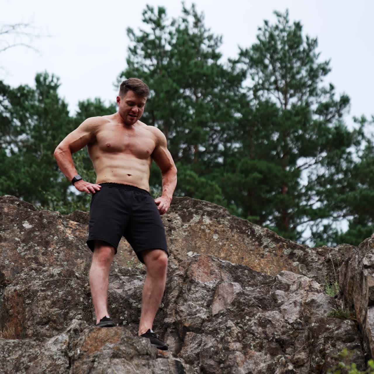 Caucasian muscular man in black shorts stand high on the rock. Sportsman demonstrates his athletic torso. Low angle view