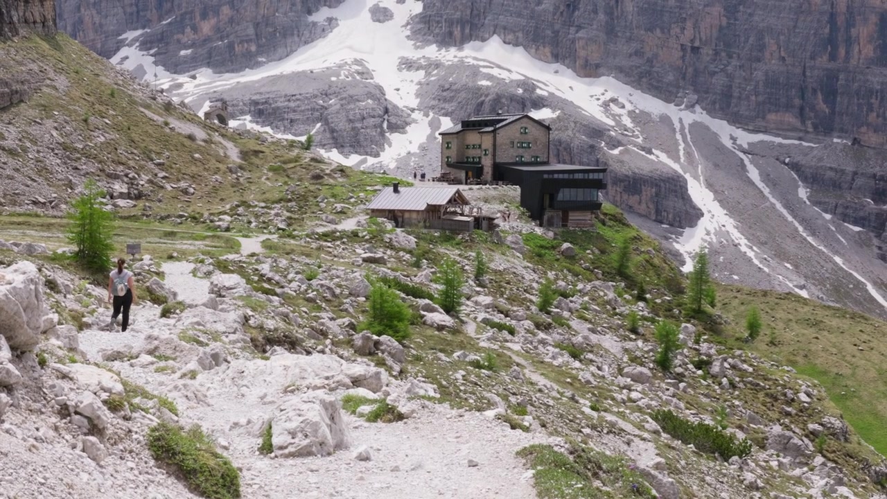 Giovane donna con zaino in spalla che fa trekking su un sentiero di montagna roccioso. Vista panoramica di un rifugio di montagna e delle cime innevate delle Dolomiti in Trentino.