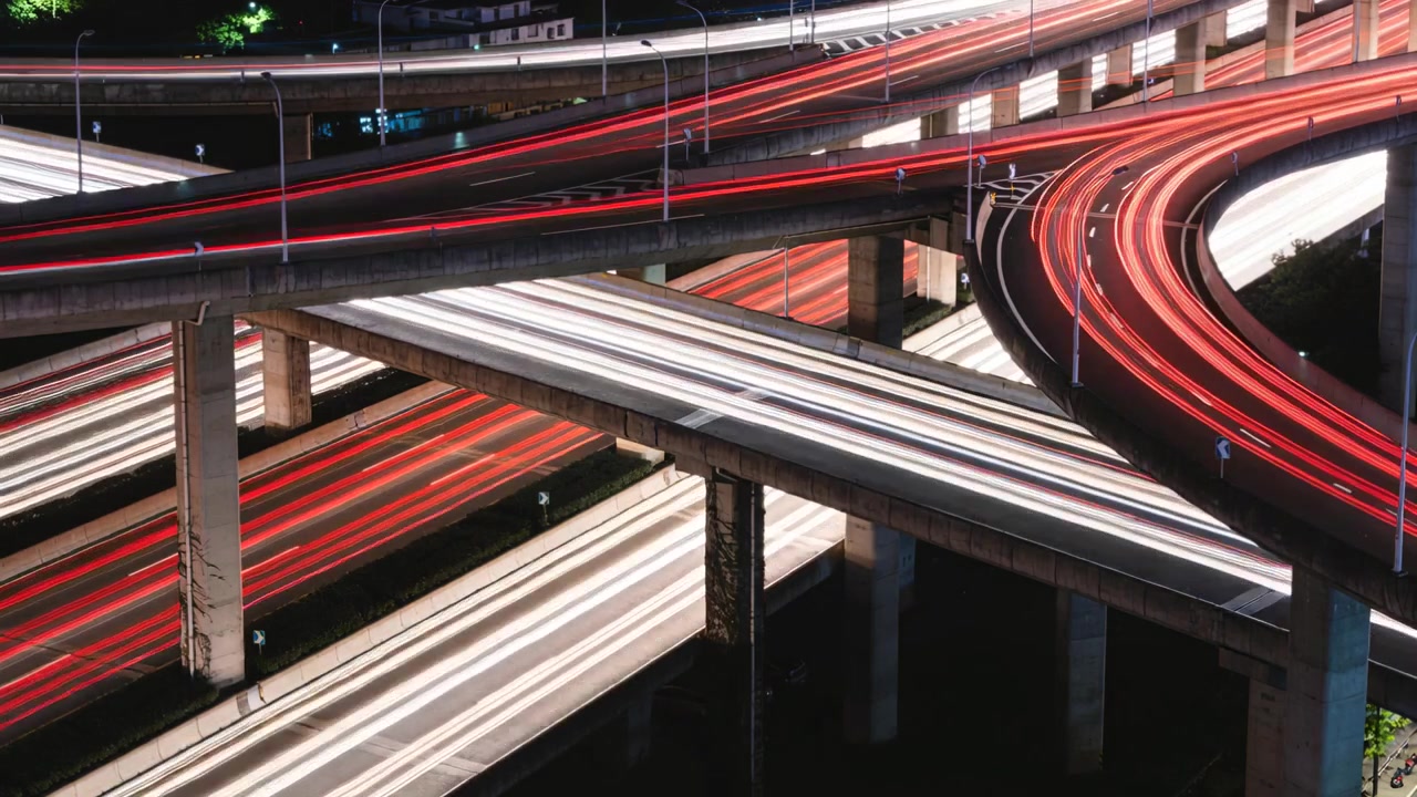 Ripresa del traffico che scorre su un cavalcavia di notte, mostrando scie luminose rosse e bianche con i piloni.