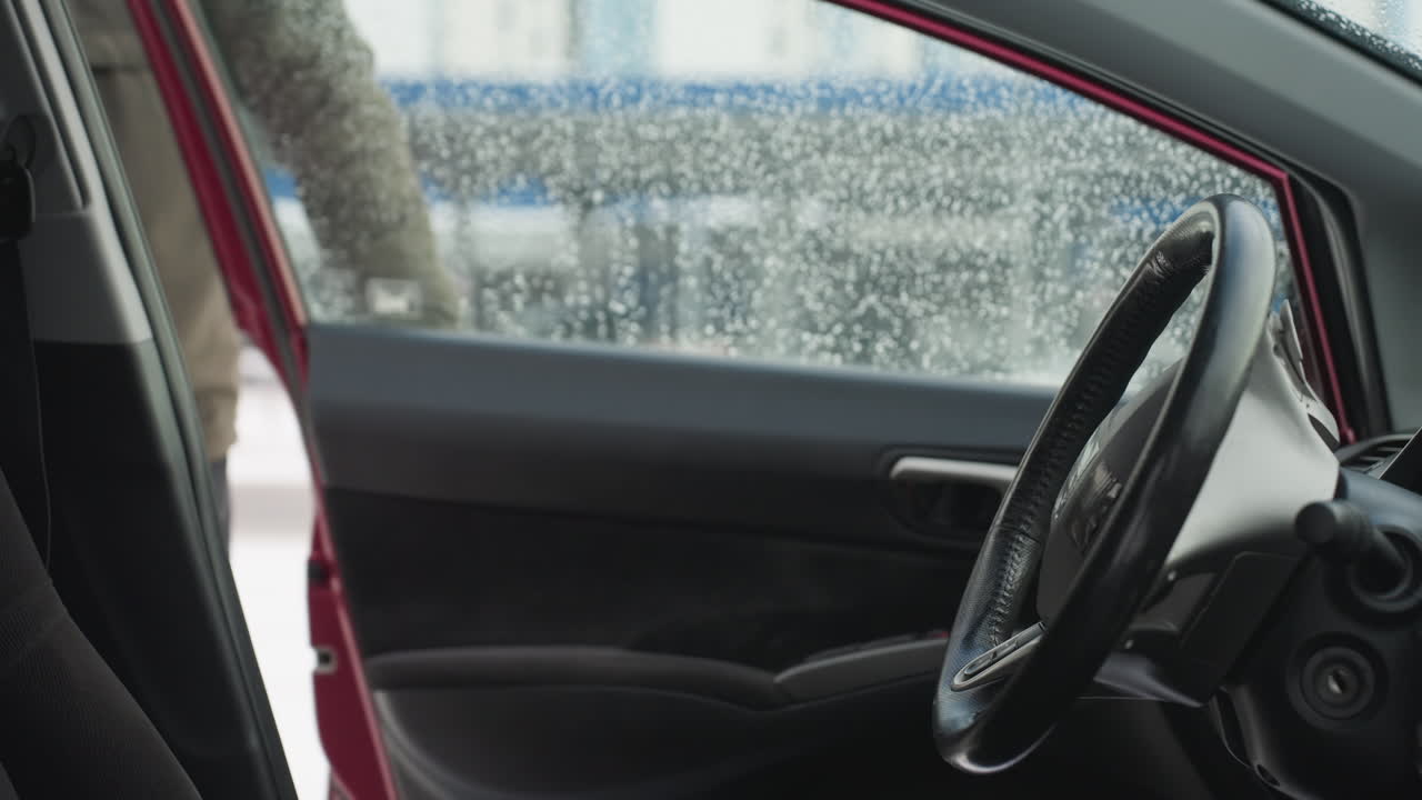 Close up from car interior as driver opens door and enters vehicle during cold weather, water droplets visible on window with urban background blurred through glass
