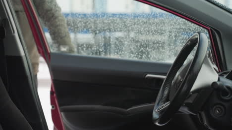 close up from car interior as driver opens door and enters vehicle during cold weather, water droplets visible on window with urban background blurred through glass