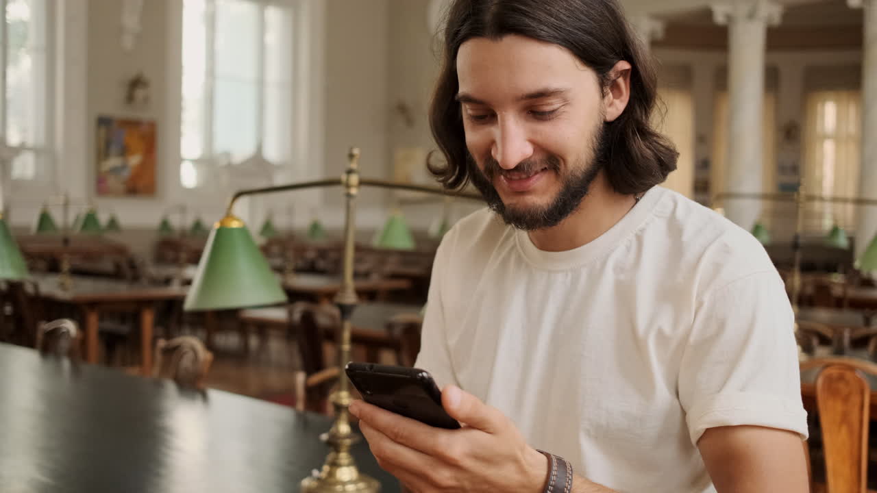 Young joyful student using cellphone in library