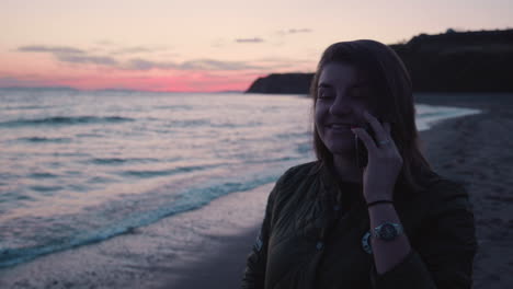 Medium-shot-of-a-happy-girl-talking-on-the-phone-while-walking-along-the-beach-on-the-sunset,-with-sea-waves-crushing-on-shore-in-the-background