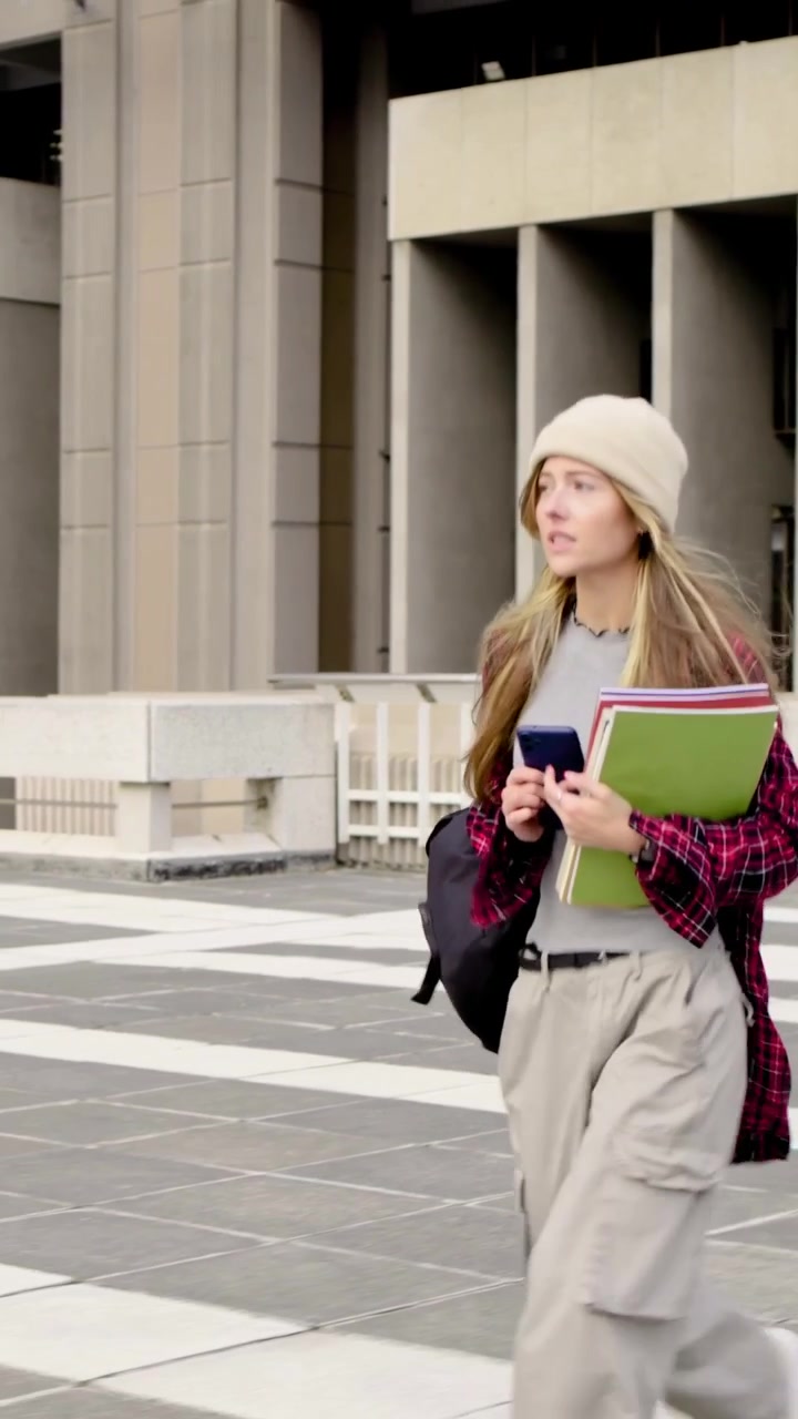 Video verticale: La telecamera segue una donna con berretto che cammina con documenti e telefono su una piazza sul tetto verso il campus.