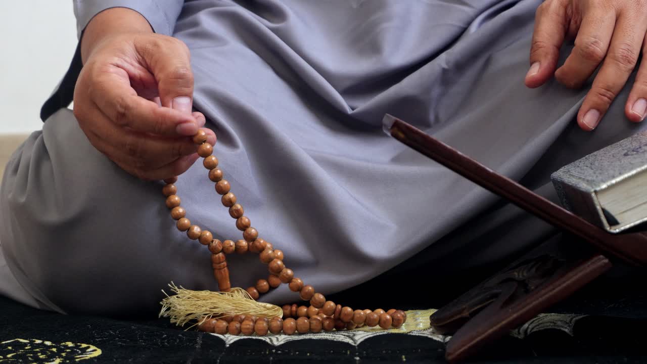 Close Up Of Man Hands On Tasbih With Small Quran On Rehal In The Background