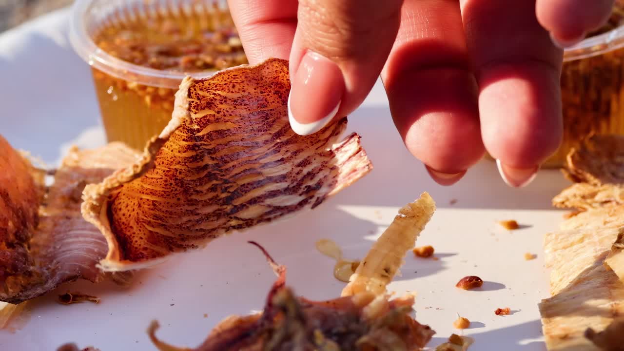 Close-up of hands selecting dried seafood pieces with a spicy dipping sauce on the side.