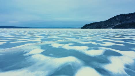 aerial footage flying low over a frozen lake with snowy patterns towards the edge of a cliff rising out of the ice