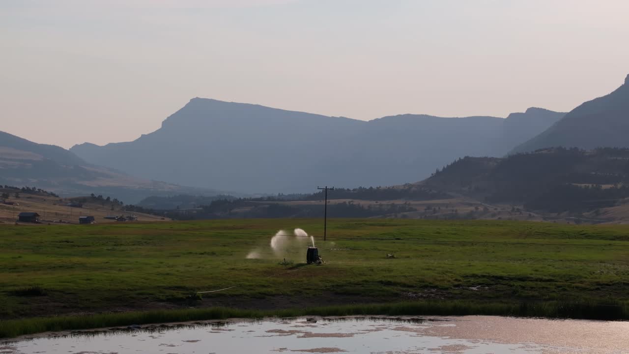 A rural field in Wyoming with sprinklers, mountains, and a pond in the early morning light