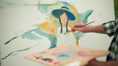 primer plano de un artista con un sombrero y una camisa a cuadros sosteniendo una paleta y pintando un retrato colorido en un lienzo. el enfoque está en la mano y el pincel mientras el artista da vida a la obra de arte al aire libre