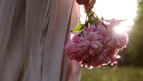 Rückseitige-Tracking-Aufnahmen-Einer-Frau-In-Heller-Kleidung,-Die-Mit-Einem-Strauß-Rosa-Blumen-In-Der-Hand-Durch-Die-Grüne-Wiese-Geht