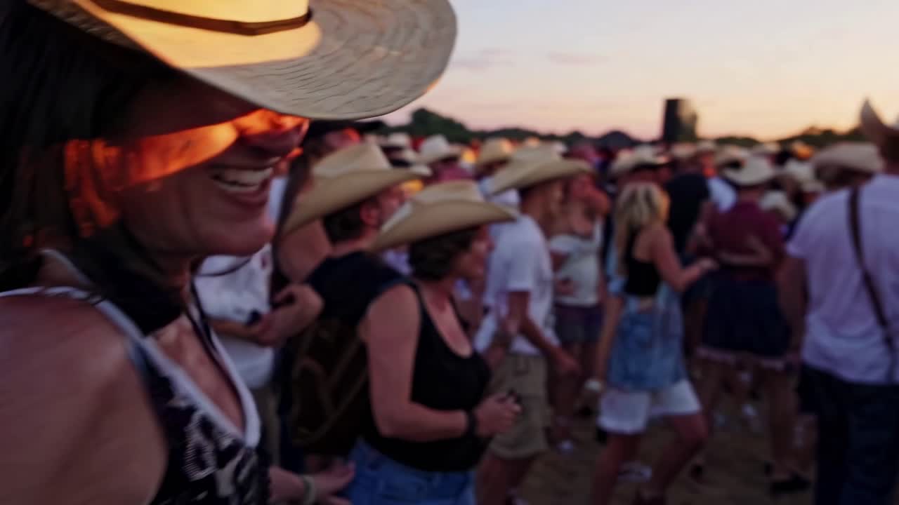 En glad kvinde med cowboyhat nyder en countrymusikfestival ved solnedgang.