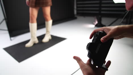 male photographer working in studio. camera in man's hands close up. lady posing in white boots at backdrop in blur.