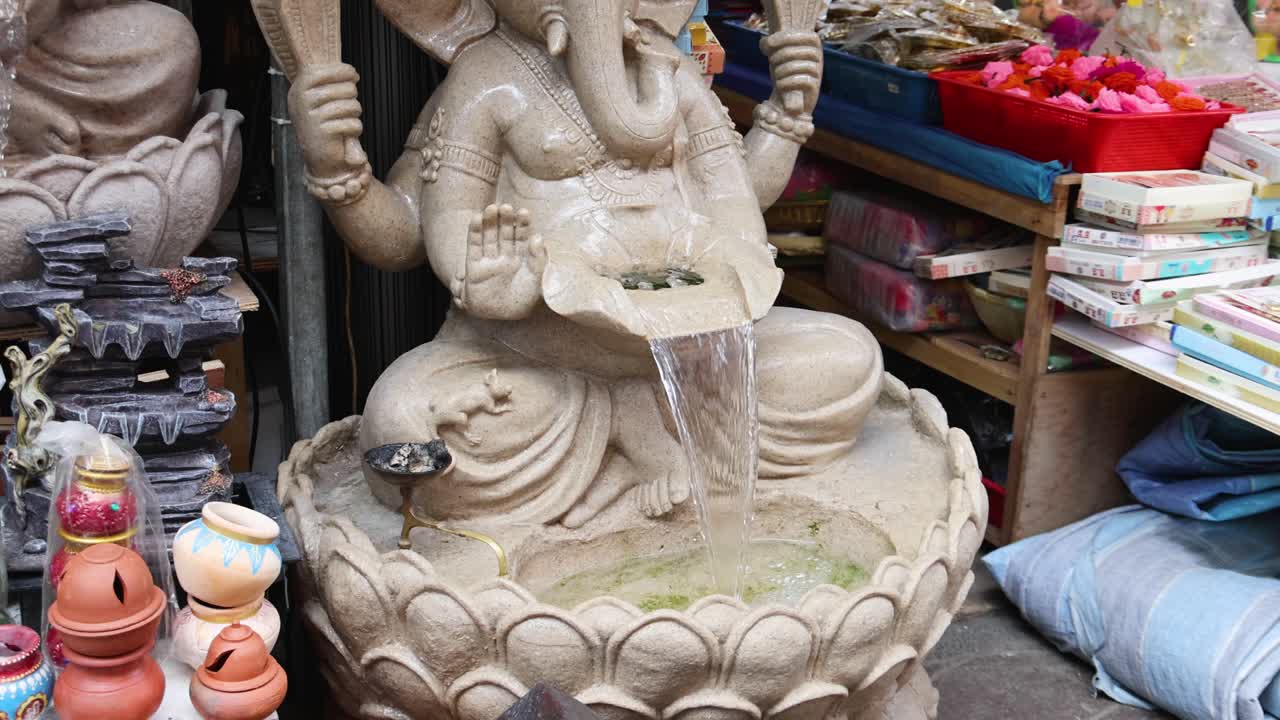 A stone Ganesha fountain continuously streams water in a colorful outdoor market, surrounded by offerings, textiles, and bright ambient daylight