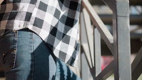 upward angle of man wearing denim jeans and checkered shirt leaning on metal railing with geometric design under sunlight, mid-step as he appears relaxed in urban setting possibly smoking