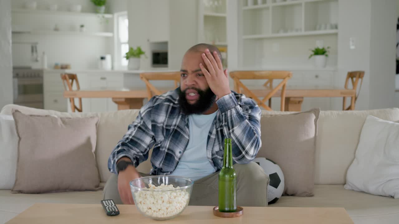 Homme afro-américain en chemise à carreaux sur le canapé regardant le football, attrapant du pop-corn, gesticulant, se frottant la tête