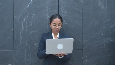 focused latin businesswoman working outside, using laptop while standing at office building wall