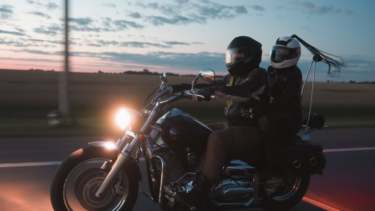 Side view of motorcycle rider and passenger on road trip during evening sunset, bright headlight glowing as couple rides across countryside with scenic sky filled with clouds