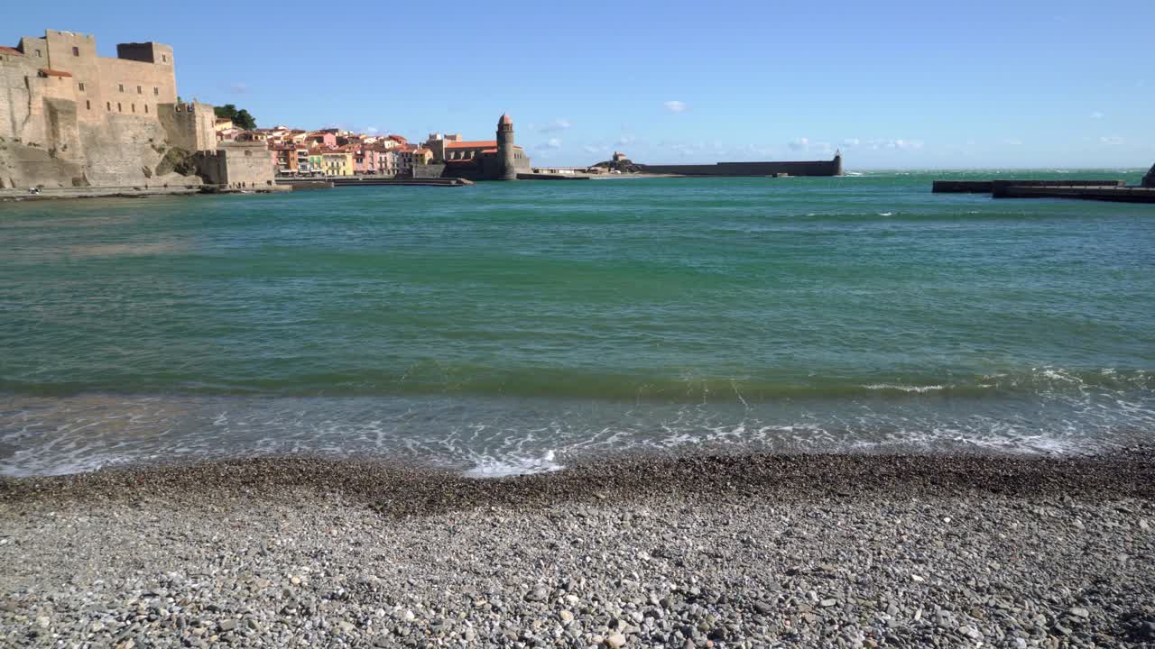 Il centro storico di Collioure visto dalla spiaggia di ciottoli, con dolci onde che lambiscono la riva in una giornata estiva ventosa.