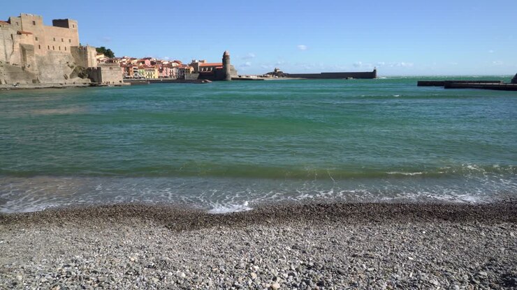 Il centro storico di Collioure visto dalla spiaggia di ciottoli, con dolci onde che lambiscono la riva in una giornata estiva ventosa.