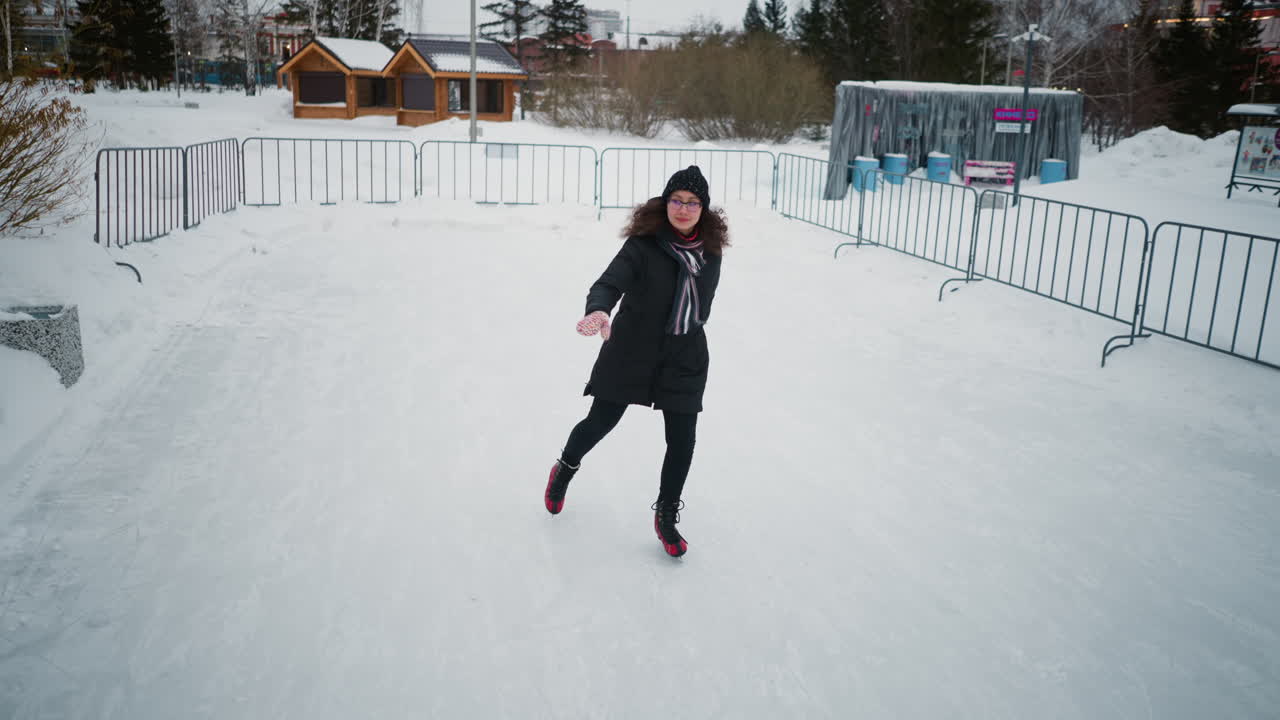 Lady skating gracefully on outdoor ice rink during winter, wearing black coat, knit hat, colorful gloves, striped scarf, and red skates, enjoying seasonal activity surrounded by snow, frosty atmosphere