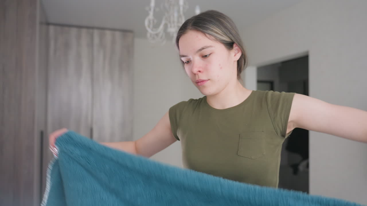 Woman arranging blue towel indoors, carefully folding soft fabric with focused expression, natural light highlighting daily household task in modern home interior with minimal background details