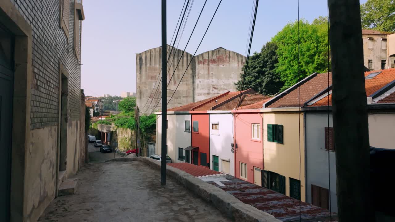 Calle estrecha de adoquines en Oporto con coloridas casas tradicionales. Un poste telefónico con numerosos cables y una farola se alzan prominentemente, conduciendo a un edificio industrial distante.