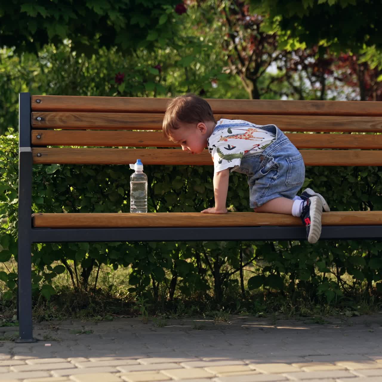 Lovely boy in a romper climbs on the wooden bench in the park. Kid tries to sit closer to the bottle of water