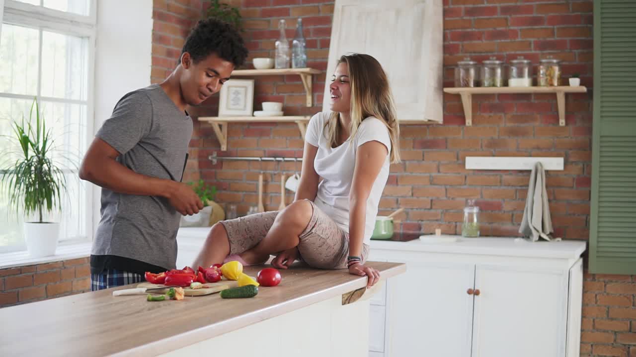 Attractive multi ethnic couple chatting in the kitchen early in the morning. Handsome man feeding his wife while cooking