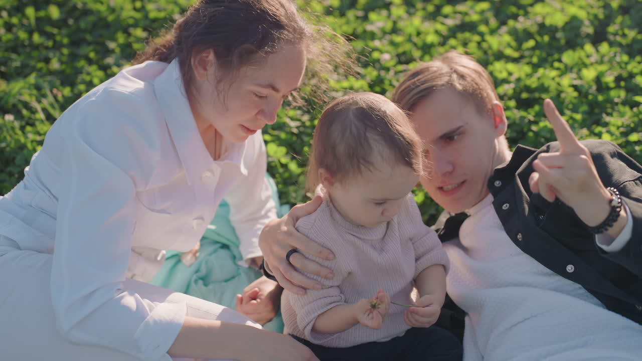 Kaukasische Eltern kuscheln mit Kleinkind auf dem Gras, frisch verheiratetes Paar genießt intimes Picknick auf sonniger Wiese, Mutter und Vater wiegen Kind mit sanftem Lächeln, weiches natürliches Licht, Nahaufnahme Zärtlichkeit.