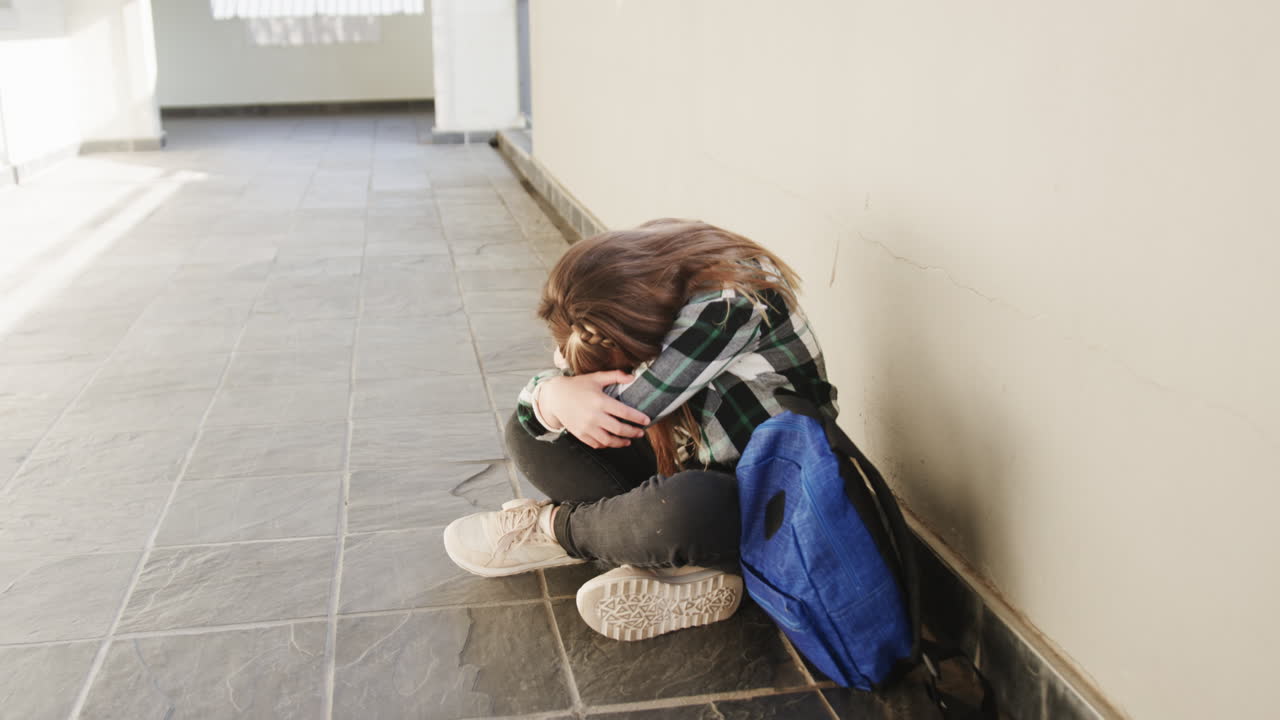 Young girl sitting in school hallway with backpack, feeling overwhelmed and stressed, copy space